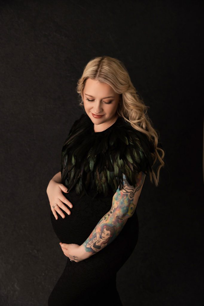 Pregnant woman dressed in black maternity dress with a feather cape around her shoulders, dark black backdrop. Mum is holding her belly while looking down at her belly with a slight smile. 
