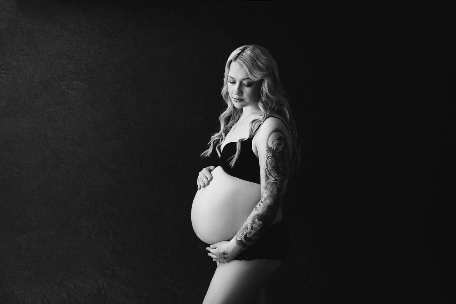 Pregnant woman dressed in black bra and underwear, dark black backdrop. Mum is holding her belly while looking down at her belly. Black and white photograph