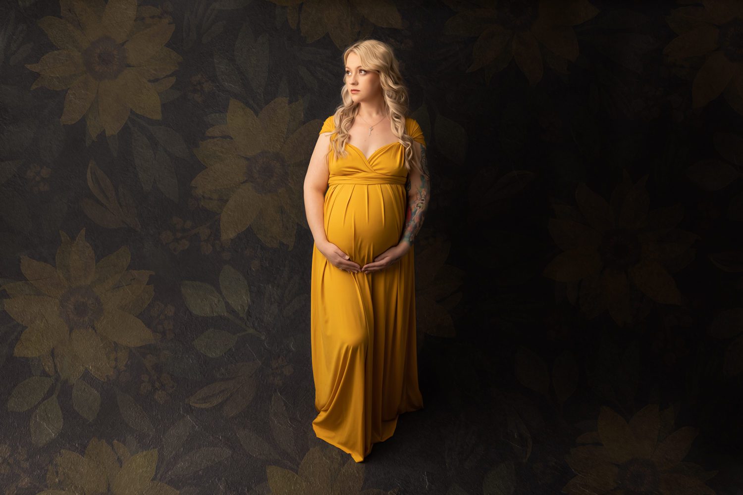 Pregnant woman dressed in yellow maternity gown, dark grey backdrop with sunflower overlay. Mum is holding her belly while looking away from the camera with a slight smile.