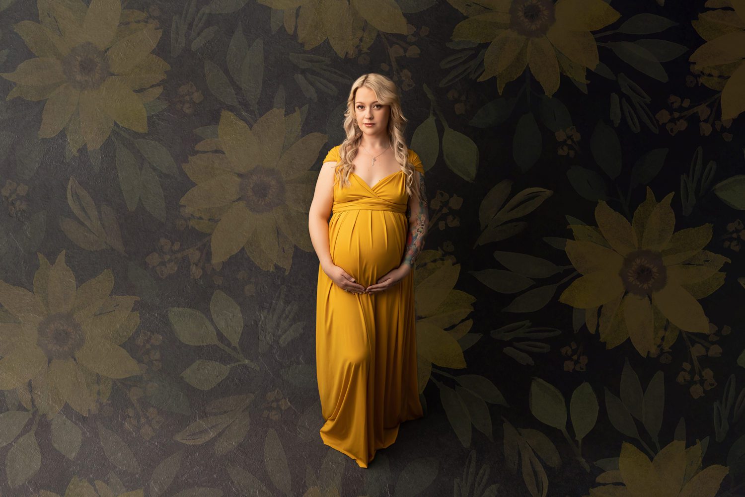 Pregnant woman dressed in yellow maternity gown, dark grey backdrop with sunflower overlay. Mum is holding her belly while looking at the camera with a slight smile.