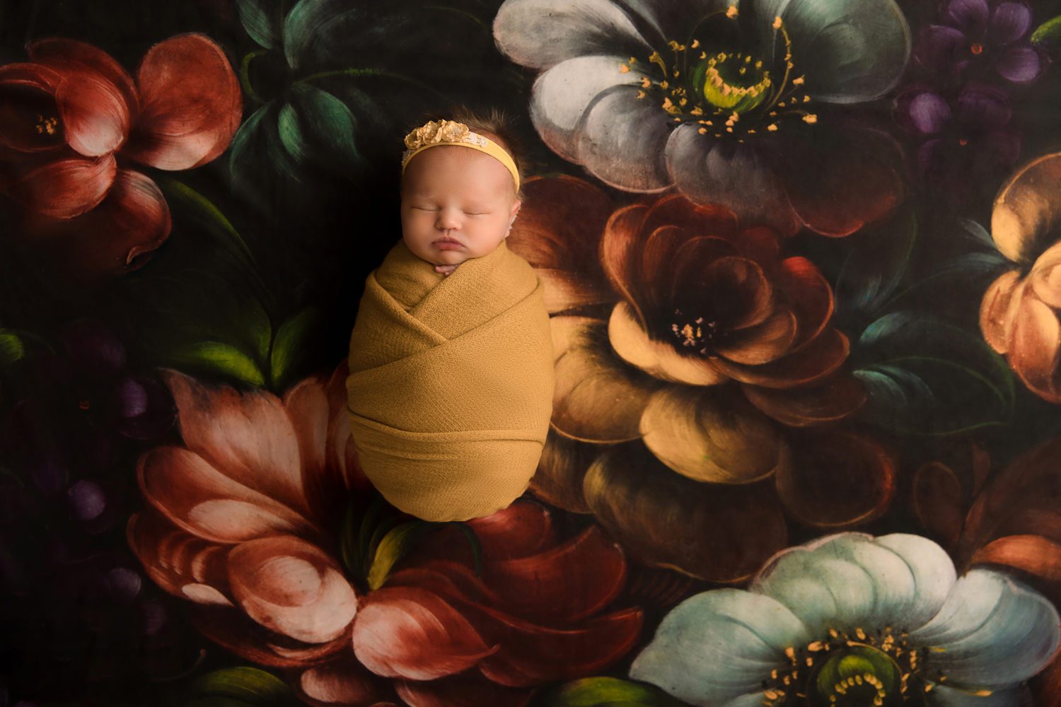 newborn baby girl wrapped in yellow wrap on dark floral backdrop