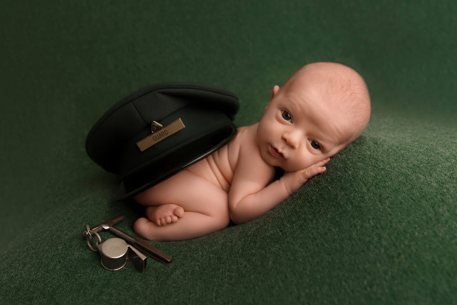 Newborn baby in bottom up pose with grandfathers hat and whistle next to him on green blanket