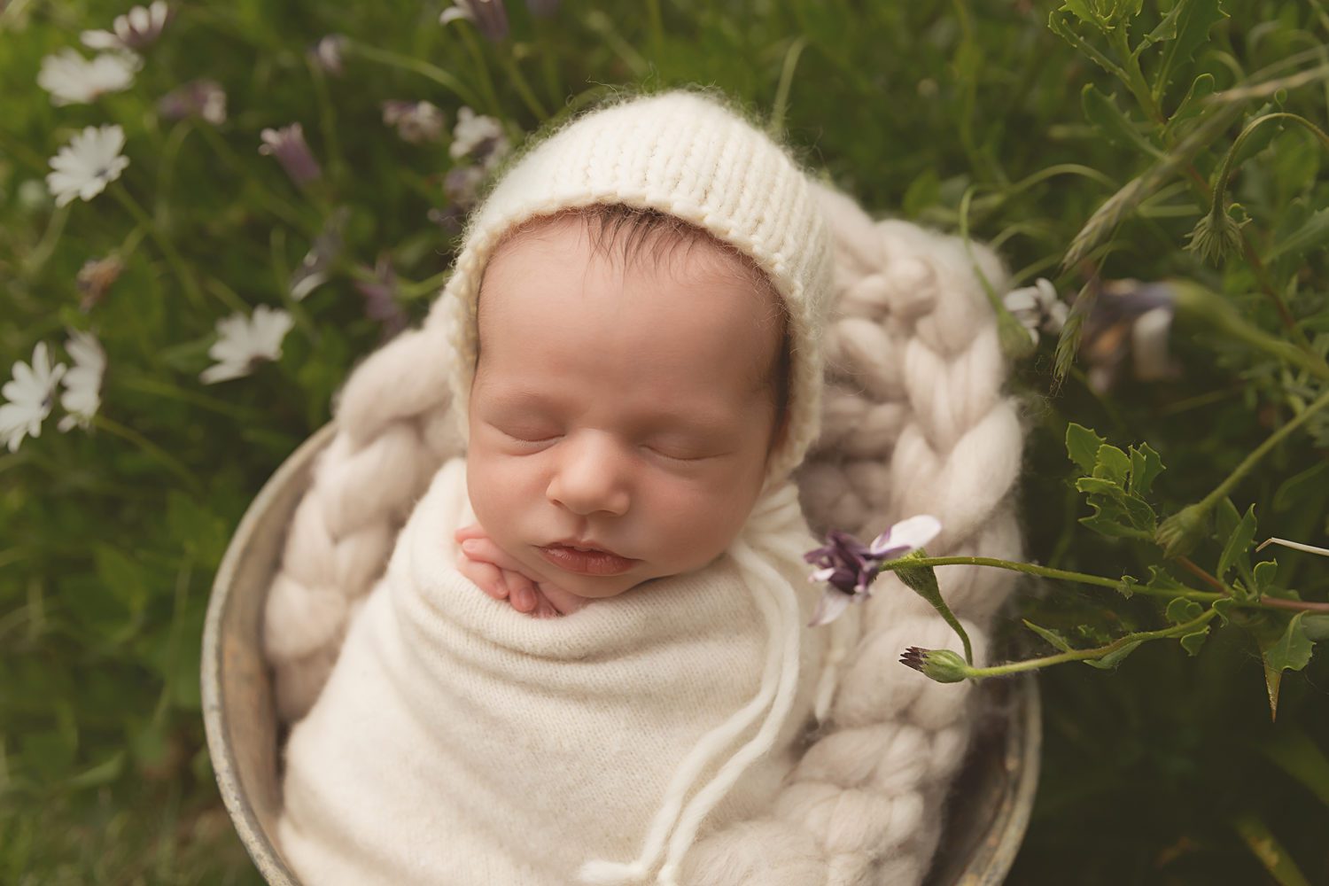 newborn baby wrapped in cream wrap in basket outside between flowers