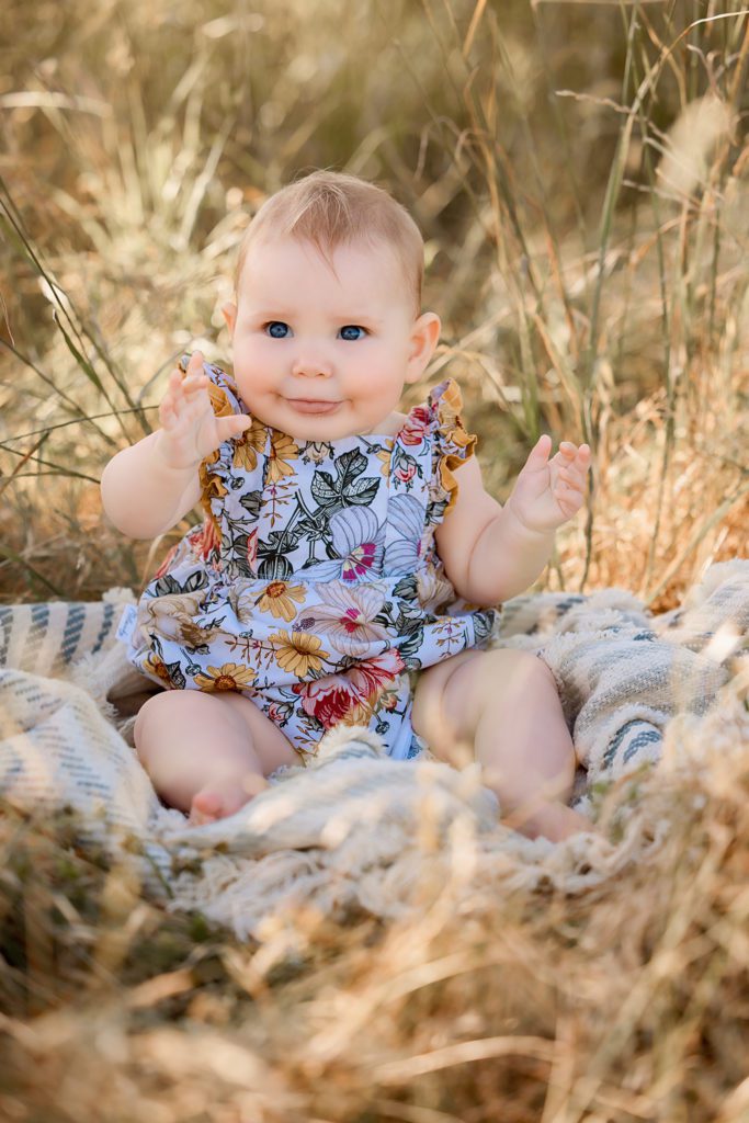Family photo baby girl under 1 year sitting on blanket in grassy field. floral outfit on. cheeky face.