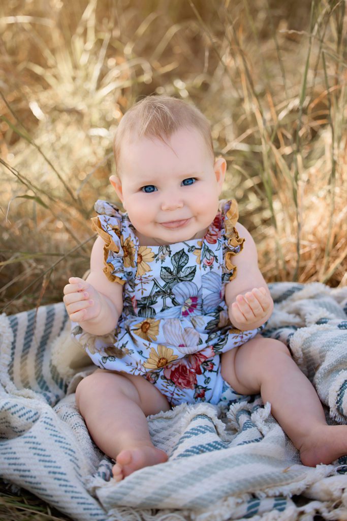 Family photo baby girl under 1 year sitting on blanket in grassy field. floral outfit on. cheeky face.