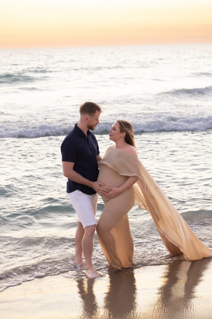 Expectant couple laughing and holding each other on Scarborough Beach with ocean waves crashing behind them — maternity photography by Aurora Joy Photography, Scarborough Western Australia Perth maternity photographer