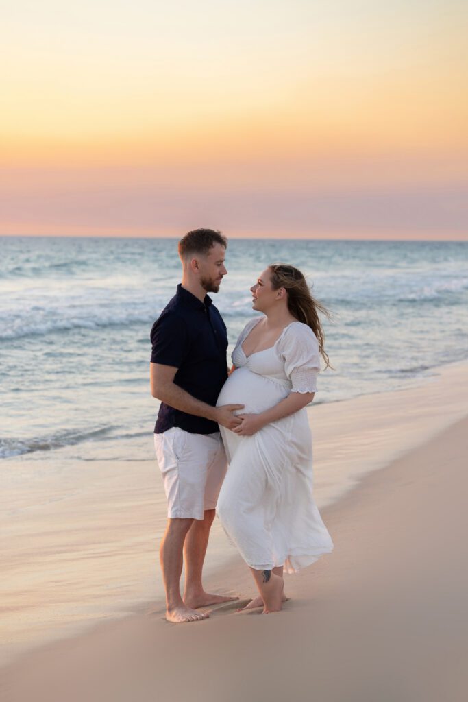 Expectant couple ,on a Perth beach mother elegantly draped in soft white fabric in a side profile pose, showcasing her baby bump — maternity photography by Aurora Joy Photography, Scarborough Western Australia