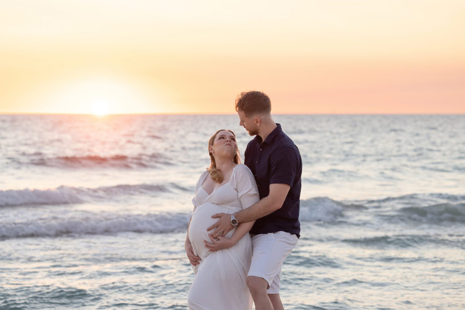 Expectant couple ,on a Perth beach mother elegantly draped in soft white fabric in a side profile pose, showcasing her baby bump — maternity photography by Aurora Joy Photography, Scarborough Western Australia Perth maternity photographer
