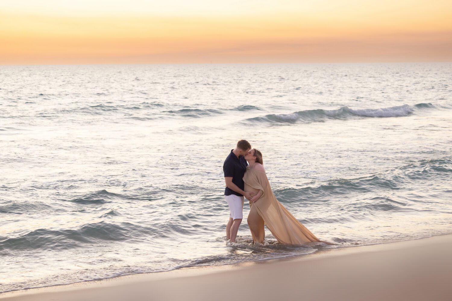 Expectant couple laughing and holding each other on Scarborough Beach with ocean waves crashing behind them — maternity photography by Aurora Joy Photography, Scarborough Western Australia