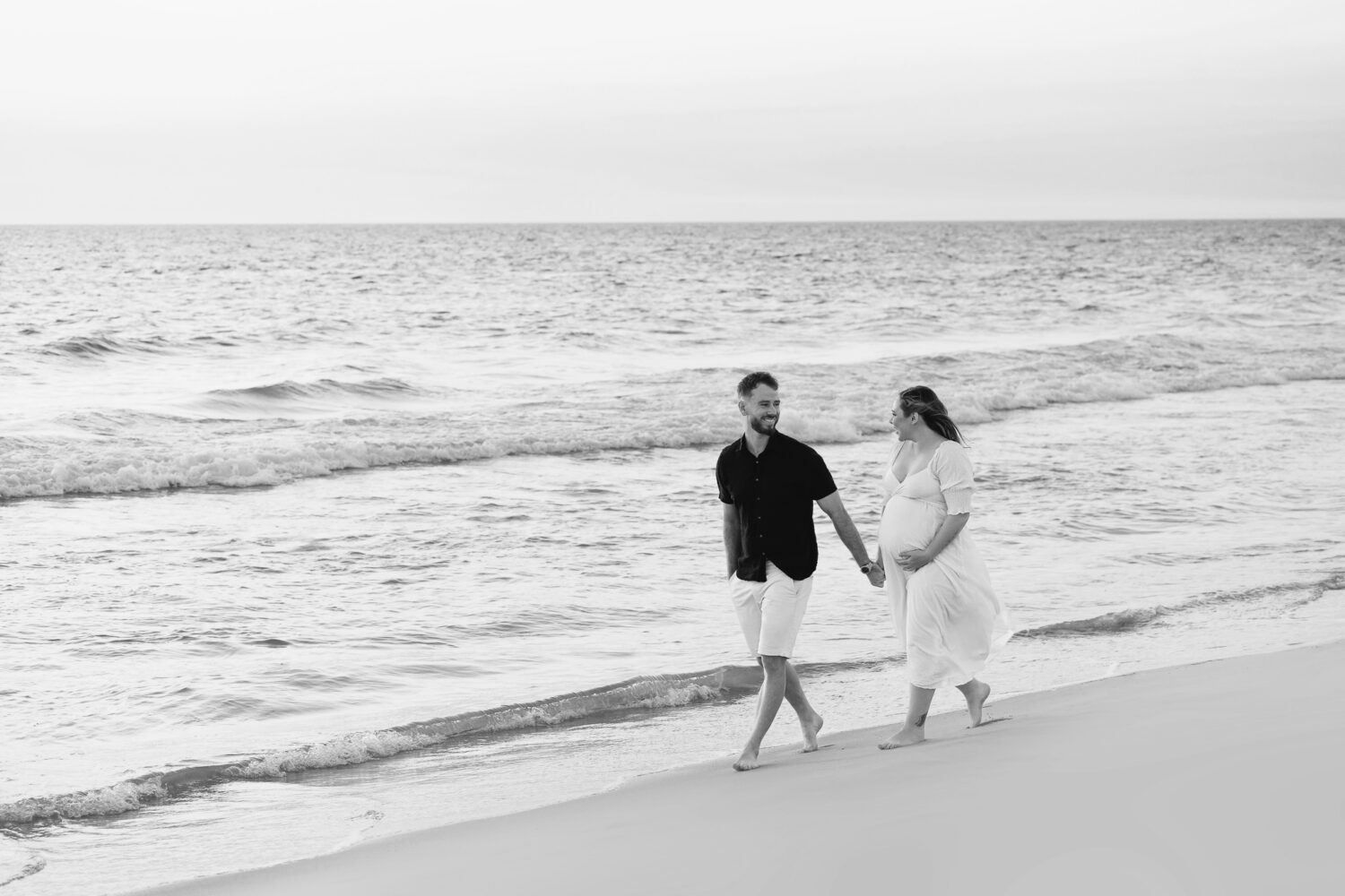 Expectant couple ,on a Perth beach mother elegantly draped in soft white fabric in a side profile pose, showcasing her baby bump — maternity photography by Aurora Joy Photography, Scarborough Western Australia