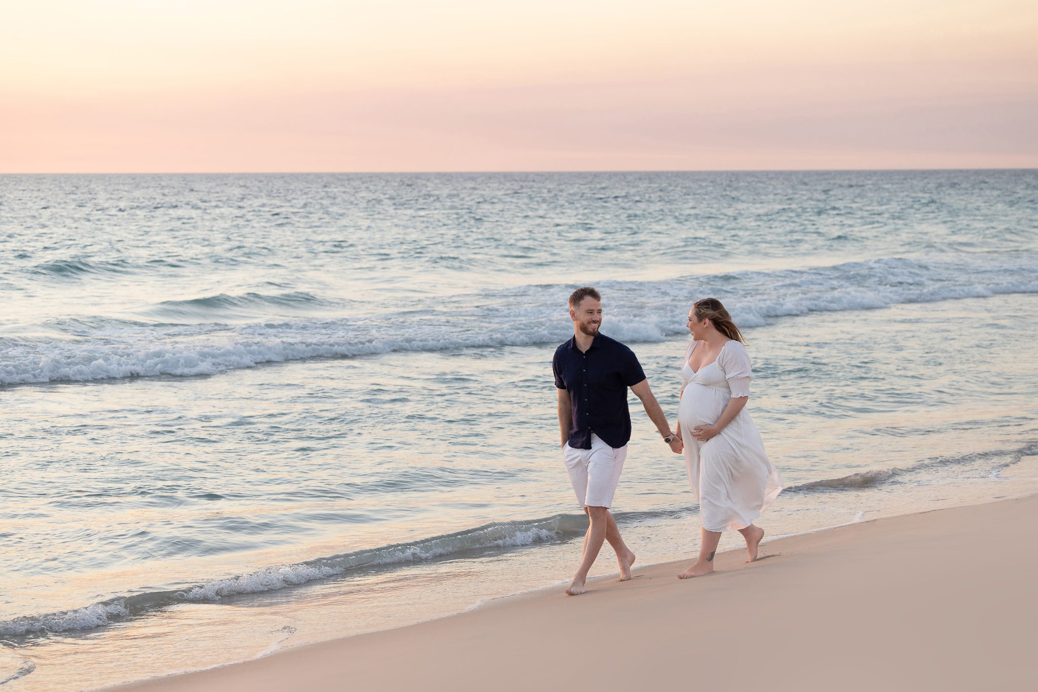 Expectant couple laughing and holding each other on Scarborough Beach with ocean waves crashing behind them — maternity photography by Aurora Joy Photography, Scarborough Western Australia