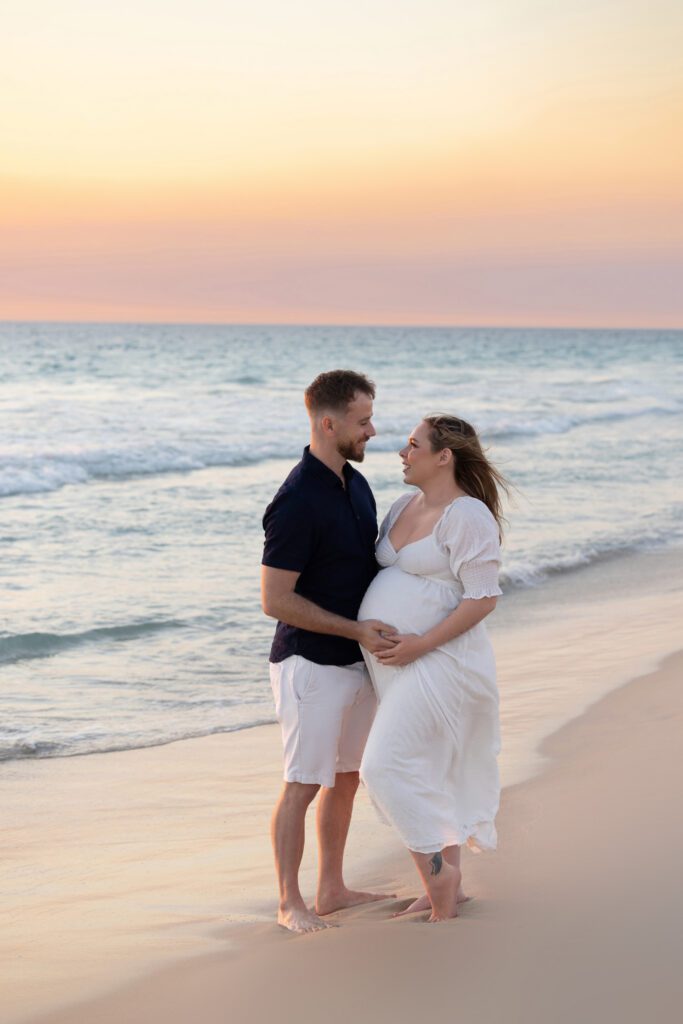 Expectant couple ,on a Perth beach mother elegantly draped in soft white fabric in a side profile pose, showcasing her baby bump — maternity photography by Aurora Joy Photography, Scarborough Western Australia