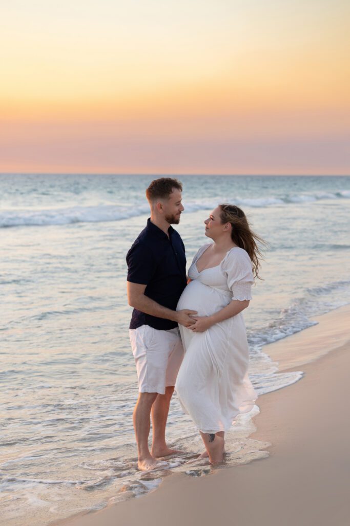 Expectant couple ,on a Perth beach mother elegantly draped in soft white fabric in a side profile pose, showcasing her baby bump — maternity photography by Aurora Joy Photography, Scarborough Western Australia